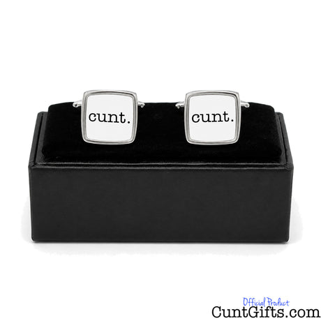 A pair of square silver cufflinks with white faces printed with the word 'cunt.' in black lowercase letters, displayed on a black presentation box against a white background.