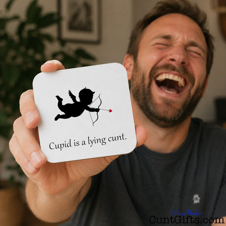 A man in a gray t-shirt is laughing while holding up a white square coaster with rounded corners. The coaster features a black silhouette of Cupid with a red arrow and the text 'Cupid is a lying cunt.