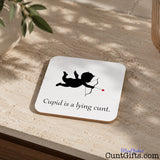 A square white coaster with rounded corners sits on a beige stone surface. The coaster has a black silhouette of Cupid with a bow and arrow and the text 'Cupid is a lying cunt'.
