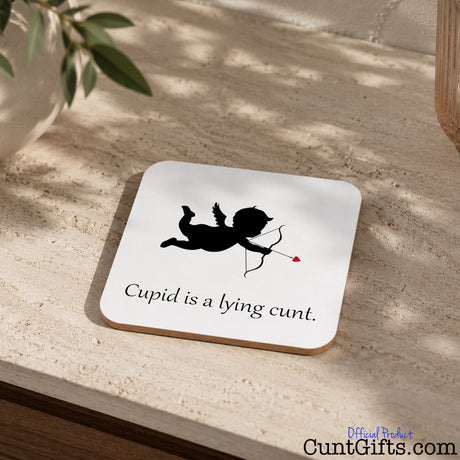 A square white coaster with rounded corners sits on a beige stone surface. The coaster has a black silhouette of Cupid with a bow and arrow and the text 'Cupid is a lying cunt'.