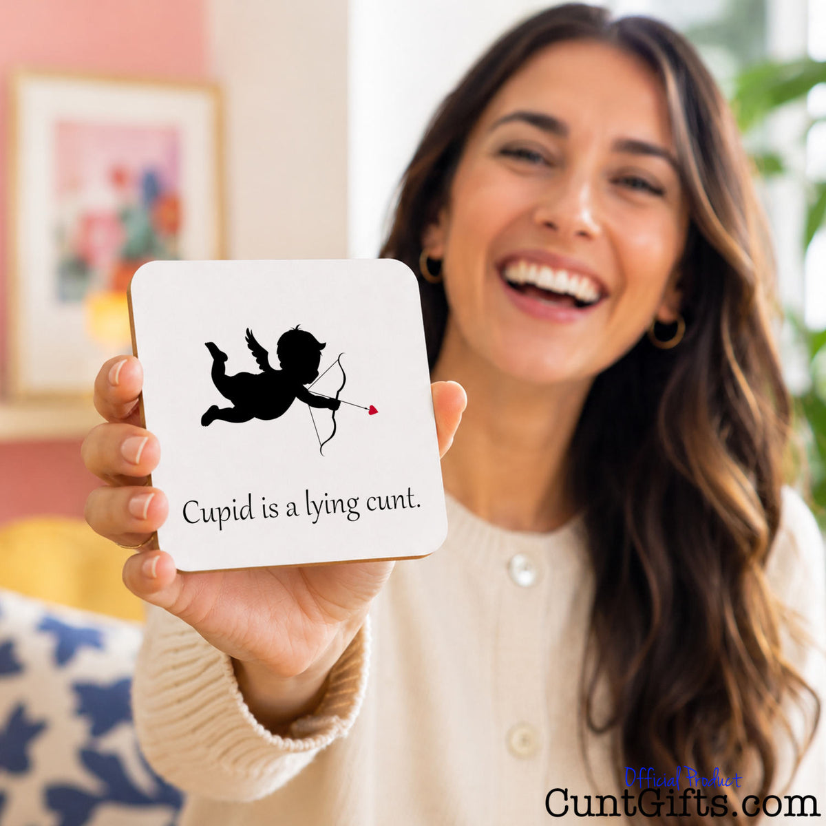 A smiling woman holds up a square white card with a black cupid silhouette and the text 'Cupid is a lying cunt.' in a home setting.