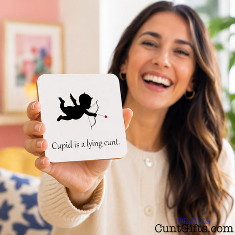 A smiling woman holds up a square white card with a black cupid silhouette and the text 'Cupid is a lying cunt.' in a home setting.