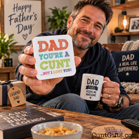 A smiling man in a cozy living room on Father's Day setting holds up a square white coaster that reads 'Dad You're A Cunt But I Love You".
