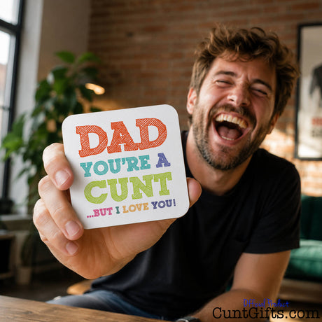 A man in a black t-shirt is laughing while holding up a square white drink coaster with colorful text that reads "Dad You're A Cunt But I Love You".
