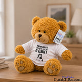 Even Jesus Think You're a Cunt teddy bear plush sitting on wooden sideboard with photo frame, plant and lamp in background.