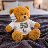 Even Jesus Think You're a Cunt teddy bear plush sitting on bed with purple throw and bedside lamp on in background.
