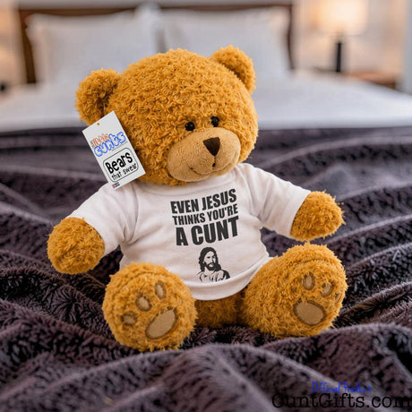 Even Jesus Think You're a Cunt teddy bear plush sitting on bed with purple throw and bedside lamp on in background.