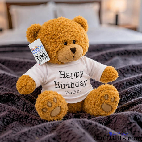 Happy Birthday You Cunt - Teddy Bear Plush sitting on purple throw on a bed.