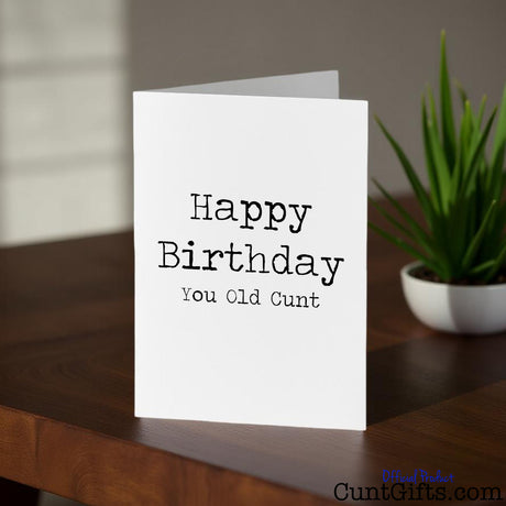 Happy Birthday You Old Cunt birthday card on a wooden surface with a plant in the background