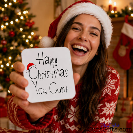 Happy Christmas You Cunt drink coaster held by laughing woman in festive hat with tree and stockings in background.