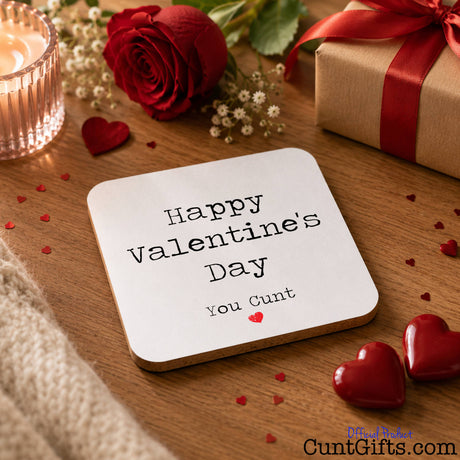 Happy Valentines Day You Cunt - drink coaster on wooden table with heart pebbles and a rose.