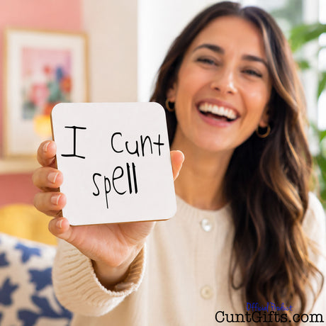 A smiling woman holds up a square white drinks coaster that reads 'I cunt spell' in black handwritten text. The background shows a softly blurred indoor room with warm tones, framed wall art, and a plant.