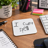 A square white drinks coaster with rounded corners on a wooden desk, featuring the handwritten black text 'I cunt spell'. The desk is styled with a keyboard, a pen, a notebook, a plant in a white pot.