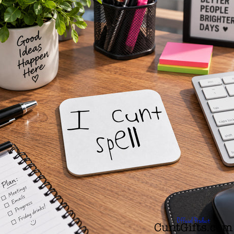A square white drinks coaster with rounded corners on a wooden desk, featuring the handwritten black text 'I cunt spell'. The desk is styled with a keyboard, a pen, a notebook, a plant in a white pot.