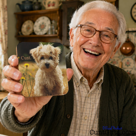 An elderly man with white hair and glasses smiles while holding up a square drinks coaster featuring a printed photo of a small fluffy dog with the word cunt. The background shows a cozy, vintage-style room with shelves of decorative plates and dishes.