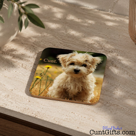 A square coaster with rounded corners featuring a printed photo of a fluffy cream-colored dog in a field with the word cunt, placed on a textured beige stone surface with soft shadows.