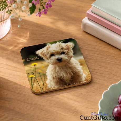 A square coaster featuring a printed photo of a small fluffy cream-colored dog sitting in a field and the word cunt, placed on a wooden tabletop with books, flowers, and a bowl nearby.