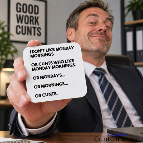 A man in a suit holds up a square white coaster with black text that reads, 'I DON'T LIKE MONDAY MORNINGS. OR CUNTS WHO LIKE MONDAY MORNINGS.
