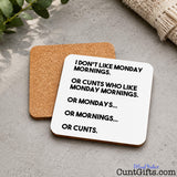 Two square cork coasters on a stone table, one showing the brown cork side and the other showing a white side with black text that reads, 'I DON'T LIKE MONDAY MORNINGS. OR CUNTS WHO LIKE MONDAY MORNINGS.