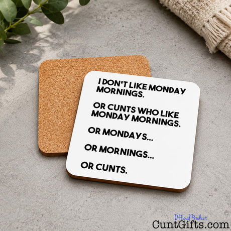 Two square cork coasters on a stone table, one showing the brown cork side and the other showing a white side with black text that reads, 'I DON'T LIKE MONDAY MORNINGS. OR CUNTS WHO LIKE MONDAY MORNINGS.
