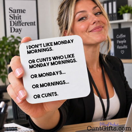 A woman in an office setting holds up a white square drinks coaster with black text that reads, 'I DON'T LIKE MONDAY MORNINGS. OR CUNTS WHO LIKE MONDAY MORNINGS.