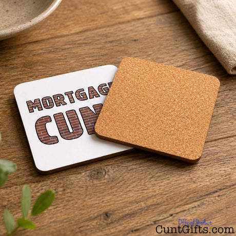 Two square coasters on a wooden tabletop. One coaster is white with the words “MORTGAGE CUNT” in a brick-style font.