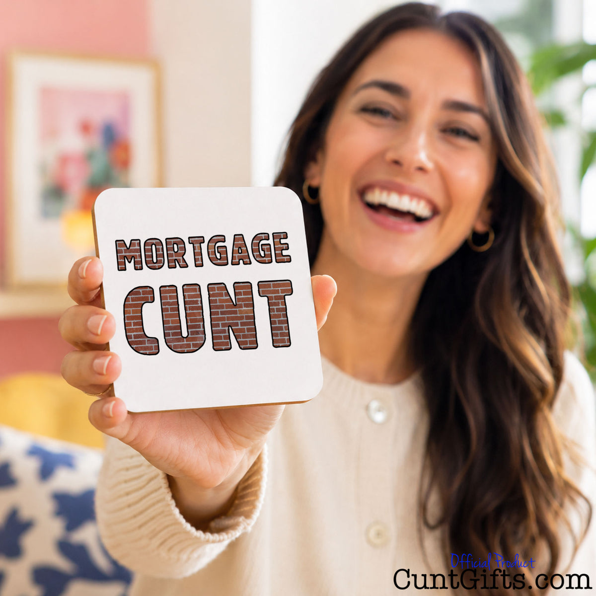 A smiling woman holds up a square white coaster with the words 'MORTGAGE CUNT' in a brown plaid font. The background is a softly blurred indoor setting with warm tones.