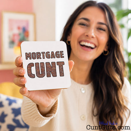 A smiling woman holds up a square white coaster with the words 'MORTGAGE CUNT' in a brown plaid font. The background is a softly blurred indoor setting with warm tones.