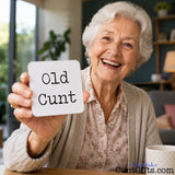 An elderly woman smiling and holding up a small white square drinks coaster that reads 'Old Cunt' in black typewriter-style text. The scene is set in a cozy indoor environment with a blurred background, and a mug is visible on the table in the foreground.