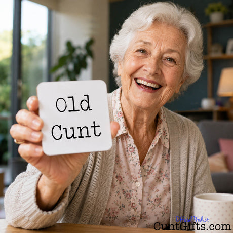 An elderly woman smiling and holding up a small white square drinks coaster that reads 'Old Cunt' in black typewriter-style text. The scene is set in a cozy indoor environment with a blurred background, and a mug is visible on the table in the foreground.