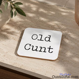A square whitedrinks coaster with rounded corners and the phrase 'Old Cunt' printed in black script, shown on a textured beige stone surface with natural shadows and plant leaves nearby.