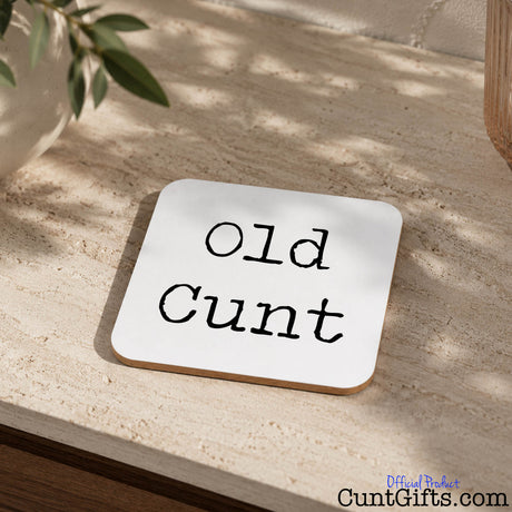 A square whitedrinks coaster with rounded corners and the phrase 'Old Cunt' printed in black script, shown on a textured beige stone surface with natural shadows and plant leaves nearby.