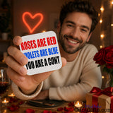 A smiling man at a romantic table setting holds up a white square drinks coaster with the text 'ROSES ARE RED VIOLETS ARE BLUE YOU ARE A CUNT' in red, blue, and black lettering. The background is warmly lit with a glowing red heart neon sign, string lights, red roses, candles, and a gift box.