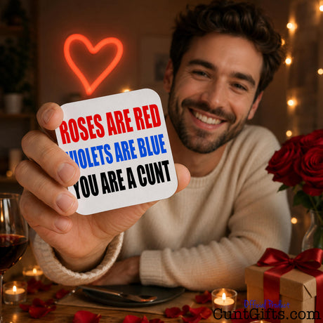 A smiling man at a romantic table setting holds up a white square drinks coaster with the text 'ROSES ARE RED VIOLETS ARE BLUE YOU ARE A CUNT' in red, blue, and black lettering. The background is warmly lit with a glowing red heart neon sign, string lights, red roses, candles, and a gift box.