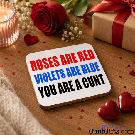 A square white drinks coaster with a wooden edge sits on a wooden table, printed with the classic nursery rhyme 'ROSES ARE RED VIOLETS ARE BLUE YOU ARE A CUNT' in red, blue, and black lettering. The scene is styled with romantic Valentine's Day props including a red rose, a lit candle, a wrapped gift box, and red heart-shaped confetti.