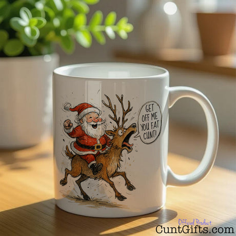 "Get Off Me You Fat Cunt" - Christmas Mug featuring Santa and Rudolph on table with plant