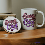 White mug and compact mirror with 'Sassy Cunt' text on a wooden surface.