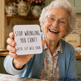 An elderly woman with white hair and glasses smiles while holding up a white square drinks coaster that reads, 'STOP WHINGING YOU CUNT! ALSO, GET WELL SOON.' in black handwritten-style text.