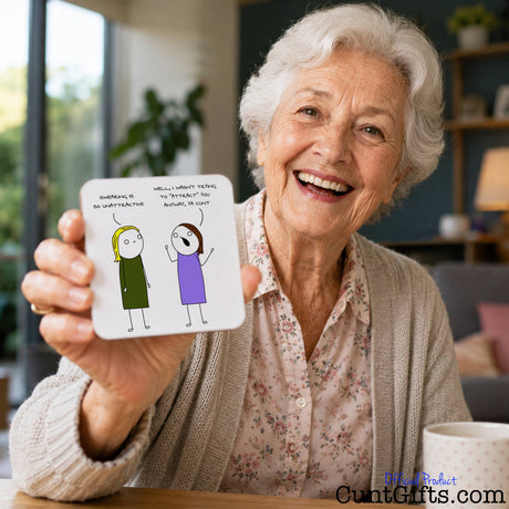 An elderly woman smiling and holding up a square white card with a simple cartoon drawing of two people funny  saying "I wasn't trying to attract you cunt" in handwritten text. and text. The scene is set in a cozy indoor environment with a window, plants, and a bookshelf in the background.