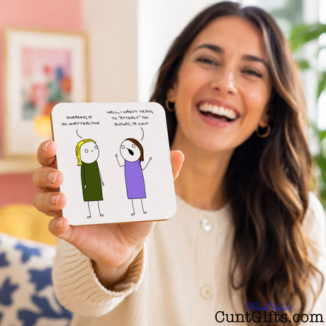 A woman smiling and holding up a square white card with a simple cartoon drawing of two figures and handwritten text and funny cartoon drawing saying "I wasn't trying to attract you cunt" in handwritten text. The scene is indoors with a softly blurred background.