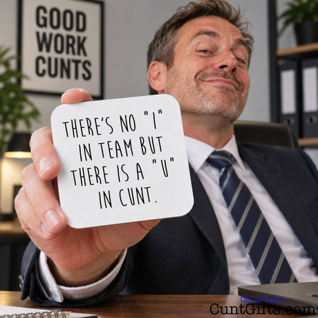 A man in a suit holding up a white square drink coaster with the text 'THERE'S NO "I" IN TEAM BUT THERE IS A "U" IN CUNT.' in a real office setting.