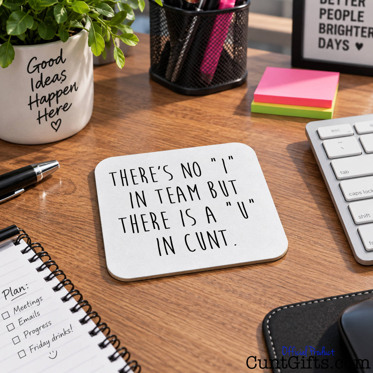 A square white coaster with black hand-lettered text reading 'THERE'S NO "I" IN TEAM BUT THERE IS A "U" IN CUNT.' displayed on a wooden desk surrounded by office items including a keyboard, notebook, plant, and sticky notes.