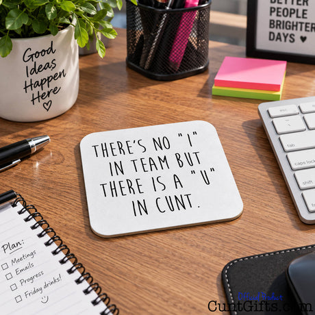 A square white coaster with black hand-lettered text reading 'THERE'S NO "I" IN TEAM BUT THERE IS A "U" IN CUNT.' displayed on a wooden desk surrounded by office items including a keyboard, notebook, plant, and sticky notes.