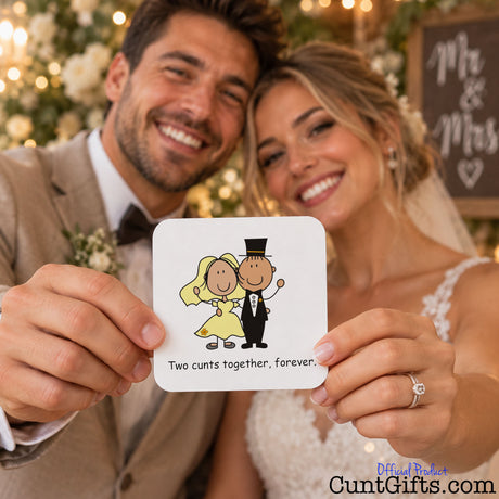 A smiling couple in wedding attire holds up a small square drink coaster with a cartoon bride-and-groom illustration and the text 'Two cunts together, forever.' The man wears a beige suit and the woman wears a white wedding dress; a blurred wedding backdrop with lights and a 'Mr.