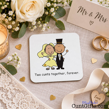 A square white wedding-themed coaster with rounded corners, featuring a cartoon bride and groom and the text 'Two cunts together, forever.' The coaster is styled on a beige fabric surface with a cream rose, white baby's breath, gold rings, a candle, lace, ribbon, and a 'Mr & Mrs' card.