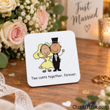 A square white wedding-themed drink coaster with rounded corners, featuring a cartoon bride and groom and the text 'Two cunts together, forever.' It is photographed on a wooden table with flowers, rings, and a blurred 'Just Married' sign in the background.