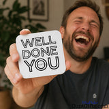 Well Done You Cunt - Drinks coaster  held by a laughing man in grey t shirt. He has a beard and is sitting in a lounge.