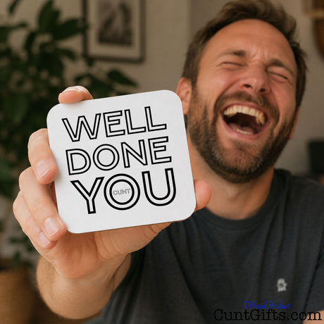 Well Done You Cunt - Drinks coaster  held by a laughing man in grey t shirt. He has a beard and is sitting in a lounge.