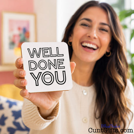 Well Done You Cunt - Drinks coaster  held by a smiling woman in white top. She is sitting in a coulorul lounge next to a plant.