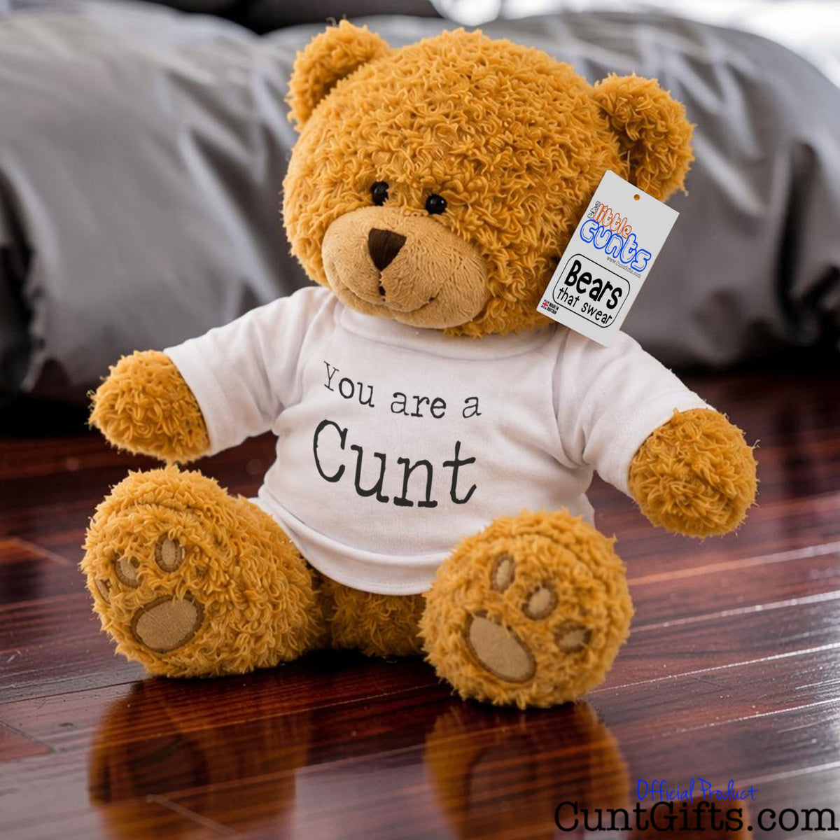 A brown plush teddy bear sitting on a wooden floor, wearing a white T-shirt that says 'You are a Cunt' in black text. The bear has a hangtag on its right ear and a blurred gray blanket in the background.